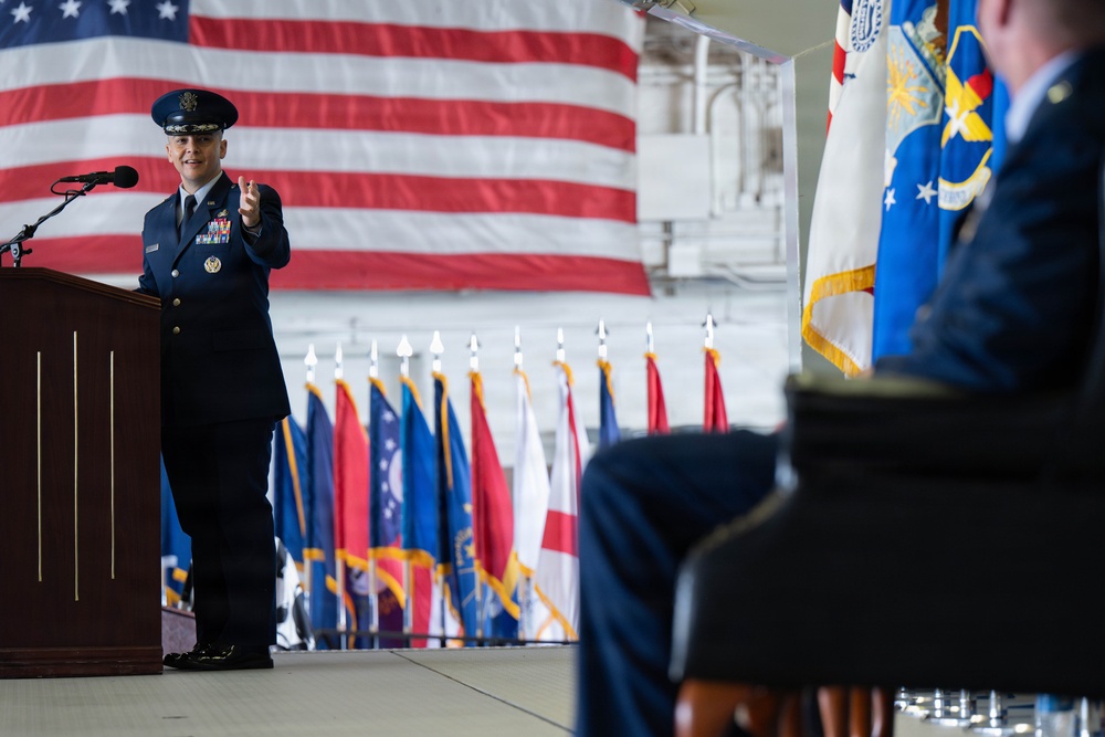 A U.S. Air Force officer in dress blues is delivering a speech at a podium during a formal military ceremony, likely a change of command or activation event. A large American flag serves as a backdrop, flanked by numerous state and military unit flags arranged in a row. A second uniformed officer is seated in the foreground to the right.