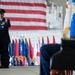 A U.S. Air Force officer in dress blues is delivering a speech at a podium during a formal military ceremony, likely a change of command or activation event. A large American flag serves as a backdrop, flanked by numerous state and military unit flags arranged in a row. A second uniformed officer is seated in the foreground to the right.