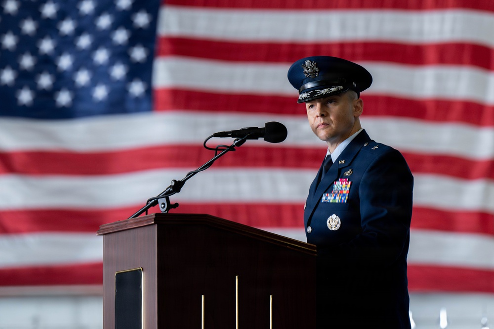 A U.S. Air Force officer in dress blues stands at a wooden podium with a microphone, positioned in front of a large American flag backdrop. The officer displays multiple service ribbons and medals, and wears a peaked cap with what appears to be general officer insignia. The setting appears to be a formal military ceremony, likely held inside a hangar or large indoor venue.