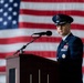 A U.S. Air Force officer in dress blues stands at a wooden podium with a microphone, positioned in front of a large American flag backdrop. The officer displays multiple service ribbons and medals, and wears a peaked cap with what appears to be general officer insignia. The setting appears to be a formal military ceremony, likely held inside a hangar or large indoor venue.
