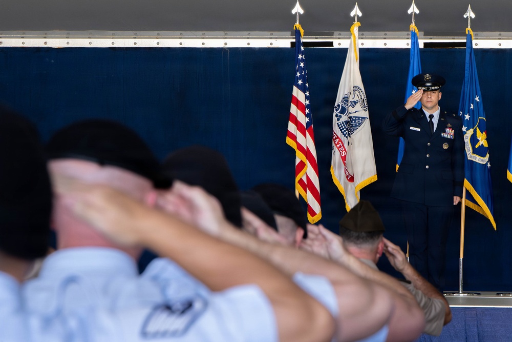 A formal U.S. Air Force military ceremony is taking place under a tent or covered venue. A uniformed Air Force officer stands at attention on a stage rendering a salute, while a group of personnel in the foreground — shown in soft focus — simultaneously salute. Three flags are displayed on stage: the U.S. flag, what appears to be a U.S. Air Force flag, and a third organizational flag.