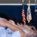 A formal U.S. Air Force military ceremony is taking place under a tent or covered venue. A uniformed Air Force officer stands at attention on a stage rendering a salute, while a group of personnel in the foreground — shown in soft focus — simultaneously salute. Three flags are displayed on stage: the U.S. flag, what appears to be a U.S. Air Force flag, and a third organizational flag.