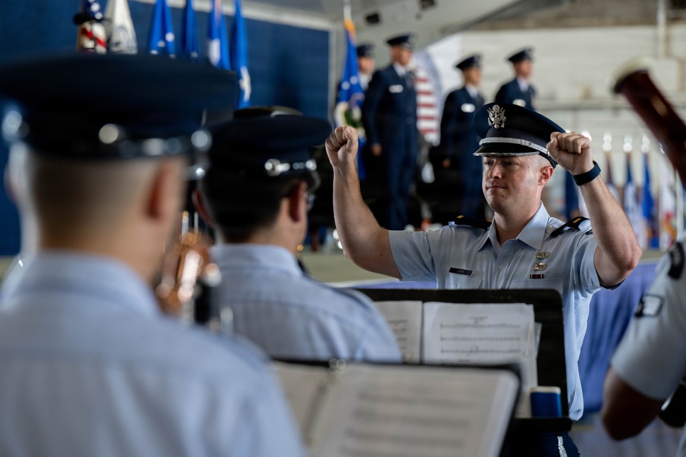 A U.S. Air Force military band conductor leads musicians during a formal ceremony, likely a change of command or official military event. The conductor, wearing a service dress uniform with an officer's cap bearing the USAF eagle insignia, has both fists raised in a conducting gesture. In the background, uniformed Air Force personnel stand at attention near multiple flags including the American flag.