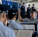 A U.S. Air Force military band conductor leads musicians during a formal ceremony, likely a change of command or official military event. The conductor, wearing a service dress uniform with an officer's cap bearing the USAF eagle insignia, has both fists raised in a conducting gesture. In the background, uniformed Air Force personnel stand at attention near multiple flags including the American flag.