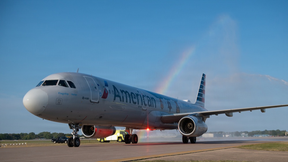 An American Airlines Airbus A321 aircraft named 'Flagship Valor' is receiving a traditional water cannon salute on an airport tarmac. The water arcs from fire trucks on both sides create a rainbow effect in the clear blue sky, indicating a ceremonial arrival or inaugural flight event. The aircraft displays American Airlines' current livery with the distinctive red, white, and blue tail design.