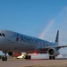 An American Airlines Airbus A321 aircraft named 'Flagship Valor' is receiving a traditional water cannon salute on an airport tarmac. The water arcs from fire trucks on both sides create a rainbow effect in the clear blue sky, indicating a ceremonial arrival or inaugural flight event. The aircraft displays American Airlines' current livery with the distinctive red, white, and blue tail design.