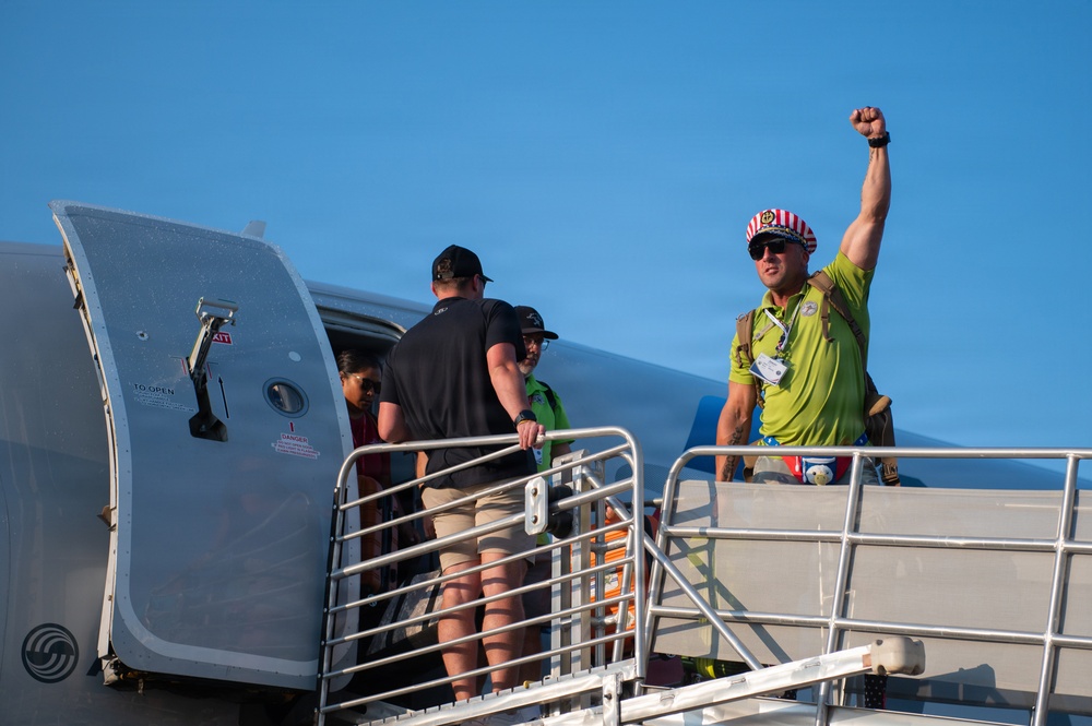 A group of people are boarding or disembarking from a large aircraft via a mobile stairway/jetway under a clear blue sky. One individual on the right, wearing a bright green shirt and a red-and-white striped hat, is raising a fist in a celebratory gesture. The scene appears to capture an arrival or departure moment with an energetic atmosphere.