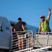 A group of people are boarding or disembarking from a large aircraft via a mobile stairway/jetway under a clear blue sky. One individual on the right, wearing a bright green shirt and a red-and-white striped hat, is raising a fist in a celebratory gesture. The scene appears to capture an arrival or departure moment with an energetic atmosphere.