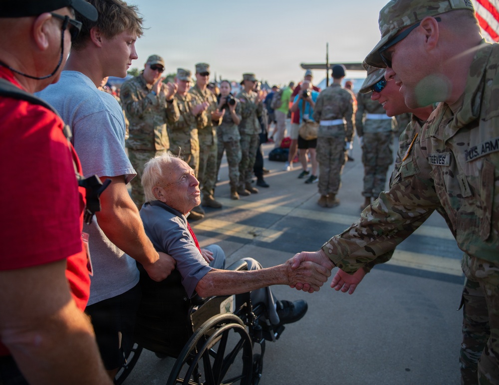 An elderly man in a wheelchair shakes hands with a U.S. Army soldier in camouflage uniform during what appears to be a military homecoming or welcome ceremony. A crowd of soldiers and civilians lines the background, many applauding. The scene conveys a moment of honor and gratitude between generations.