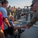 An elderly man in a wheelchair shakes hands with a U.S. Army soldier in camouflage uniform during what appears to be a military homecoming or welcome ceremony. A crowd of soldiers and civilians lines the background, many applauding. The scene conveys a moment of honor and gratitude between generations.
