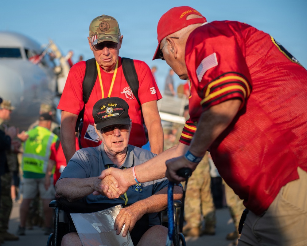 A U.S. Navy Vietnam Veteran seated in a wheelchair is being greeted with a handshake by another individual wearing a red jersey with an American flag patch. The scene appears to take place on an airport tarmac near a commercial aircraft, suggesting a honor flight or veteran recognition event. Multiple people in red shirts and yellow safety vests are visible in the background.