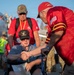 A U.S. Navy Vietnam Veteran seated in a wheelchair is being greeted with a handshake by another individual wearing a red jersey with an American flag patch. The scene appears to take place on an airport tarmac near a commercial aircraft, suggesting a honor flight or veteran recognition event. Multiple people in red shirts and yellow safety vests are visible in the background.