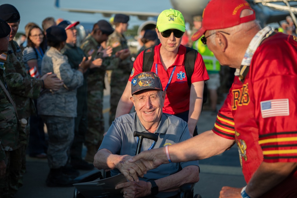 An outdoor ceremony or welcome event appears to be taking place on what looks like an airport tarmac or airfield. An elderly man in a wheelchair is shaking hands with another person wearing a red Vietnam veteran cap and jersey with an American flag patch. Military personnel in camouflage uniforms and civilians are visible in the background, suggesting this may be an honor flight or veteran recognition event.
