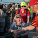 An outdoor ceremony or welcome event appears to be taking place on what looks like an airport tarmac or airfield. An elderly man in a wheelchair is shaking hands with another person wearing a red Vietnam veteran cap and jersey with an American flag patch. Military personnel in camouflage uniforms and civilians are visible in the background, suggesting this may be an honor flight or veteran recognition event.