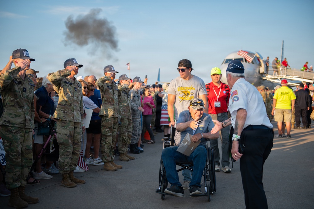 A group of military personnel in camouflage uniforms are saluting a veteran in a wheelchair being escorted along a tarmac, likely at an airport or airfield. The scene appears to be part of an honor flight or veteran recognition ceremony, with a large crowd of civilians and service members lining the path. An aircraft with an American flag is visible in the background.