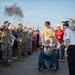 A group of military personnel in camouflage uniforms are saluting a veteran in a wheelchair being escorted along a tarmac, likely at an airport or airfield. The scene appears to be part of an honor flight or veteran recognition ceremony, with a large crowd of civilians and service members lining the path. An aircraft with an American flag is visible in the background.