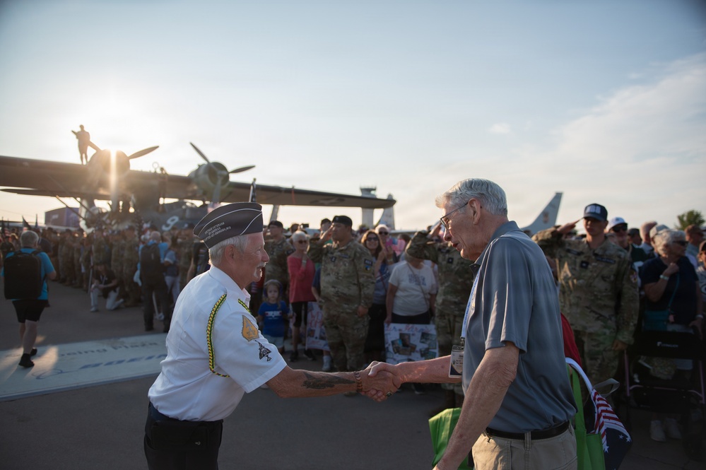 Two elderly men are shaking hands at what appears to be a military air show or veteran recognition event. One man is wearing a white veteran's uniform with a military cap and insignia, while the other is in casual civilian clothing. In the background, military personnel in camouflage uniforms are saluting, and a large crowd has gathered near vintage/historic aircraft.