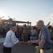 Two elderly men are shaking hands at what appears to be a military air show or veteran recognition event. One man is wearing a white veteran's uniform with a military cap and insignia, while the other is in casual civilian clothing. In the background, military personnel in camouflage uniforms are saluting, and a large crowd has gathered near vintage/historic aircraft.
