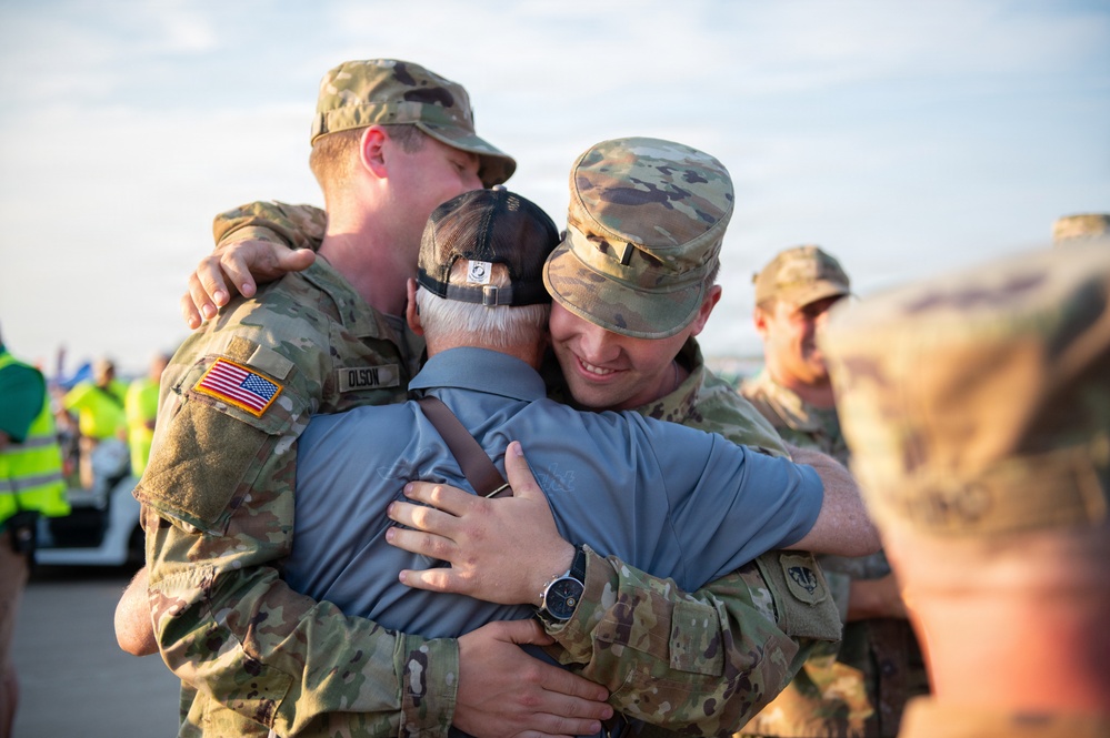 Two U.S. Army soldiers in Operational Camouflage Pattern (OCP) uniforms embrace a civilian man wearing a POW/MIA cap and blue shirt in what appears to be a homecoming or farewell event on a tarmac. The soldier on the left has a name tape reading 'OLSON' and displays a U.S. flag patch on the right shoulder. The emotional group hug suggests a reunion or deployment send-off ceremony.