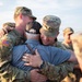 Two U.S. Army soldiers in Operational Camouflage Pattern (OCP) uniforms embrace a civilian man wearing a POW/MIA cap and blue shirt in what appears to be a homecoming or farewell event on a tarmac. The soldier on the left has a name tape reading 'OLSON' and displays a U.S. flag patch on the right shoulder. The emotional group hug suggests a reunion or deployment send-off ceremony.