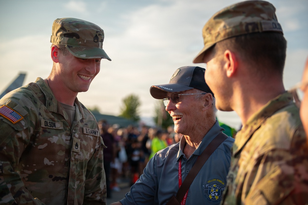 Two U.S. Army soldiers in OCP (Operational Camouflage Pattern) uniforms are engaged in a warm, smiling conversation with an elderly veteran at what appears to be an outdoor military or community event. The soldier on the left is identified by his name tape as 'OLSON' and wears Staff Sergeant rank insignia. The elderly man in the center wears a 'VETERAN' cap and a 'Yellow Ribbon Honor Flight' polo shirt, suggesting this may be an Honor Flight event.