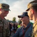 Two U.S. Army soldiers in OCP (Operational Camouflage Pattern) uniforms are engaged in a warm, smiling conversation with an elderly veteran at what appears to be an outdoor military or community event. The soldier on the left is identified by his name tape as 'OLSON' and wears Staff Sergeant rank insignia. The elderly man in the center wears a 'VETERAN' cap and a 'Yellow Ribbon Honor Flight' polo shirt, suggesting this may be an Honor Flight event.