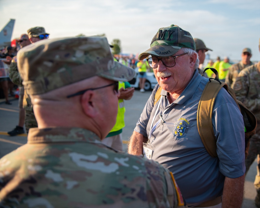 An older civilian veteran wearing an 'ARMY' camouflage cap and a gray polo shirt with a lanyard/badge is engaged in conversation with an active-duty soldier in OCP (Operational Camouflage Pattern) uniform. The setting appears to be an outdoor military or airshow event, likely a veteran recognition or honor flight program based on the visible logo on the civilian's shirt. A crowd of military personnel and civilians is visible in the background near what appears to be an aircraft.