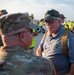 An older civilian veteran wearing an 'ARMY' camouflage cap and a gray polo shirt with a lanyard/badge is engaged in conversation with an active-duty soldier in OCP (Operational Camouflage Pattern) uniform. The setting appears to be an outdoor military or airshow event, likely a veteran recognition or honor flight program based on the visible logo on the civilian's shirt. A crowd of military personnel and civilians is visible in the background near what appears to be an aircraft.