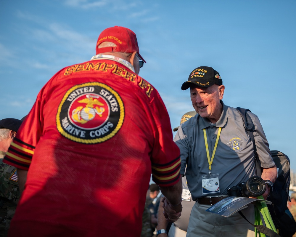 Two older men, both Vietnam veterans, are shaking hands at what appears to be a veteran's event or ceremony. The man facing away wears a red 'Semper Fi' jersey with a United States Marine Corps emblem on the back, while the man facing the camera wears a black 'Vietnam Veteran' cap and a gray polo shirt with a name badge reading 'Mike' and a yellow lanyard. The setting appears to be an outdoor daytime event with other attendees visible in the background.