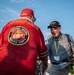 Two older men, both Vietnam veterans, are shaking hands at what appears to be a veteran's event or ceremony. The man facing away wears a red 'Semper Fi' jersey with a United States Marine Corps emblem on the back, while the man facing the camera wears a black 'Vietnam Veteran' cap and a gray polo shirt with a name badge reading 'Mike' and a yellow lanyard. The setting appears to be an outdoor daytime event with other attendees visible in the background.