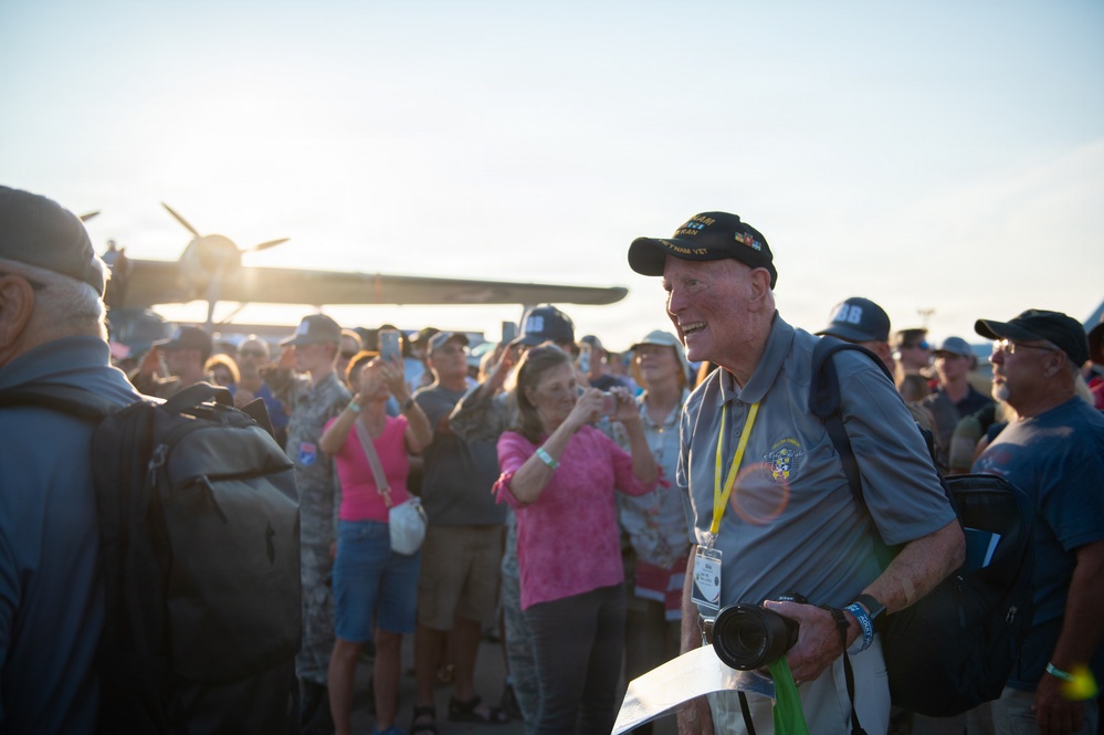 An elderly man wearing a black veteran's cap and gray shirt with a yellow lanyard stands smiling among a crowd at what appears to be an airshow or aviation event. A vintage propeller aircraft is visible in the background, backlit by bright sunlight. The crowd behind him includes people taking photographs, suggesting this may be a commemorative or special recognition event.