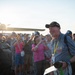An elderly man wearing a black veteran's cap and gray shirt with a yellow lanyard stands smiling among a crowd at what appears to be an airshow or aviation event. A vintage propeller aircraft is visible in the background, backlit by bright sunlight. The crowd behind him includes people taking photographs, suggesting this may be a commemorative or special recognition event.