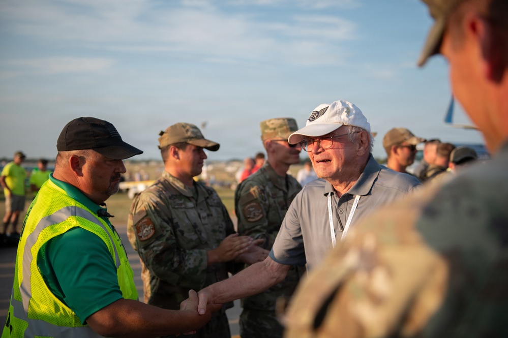 An outdoor gathering at what appears to be an airfield or aviation event, where a civilian elderly man in a white cap and gray polo shirt is shaking hands with a person wearing a high-visibility safety vest. Several military personnel in camouflage uniforms are present in the background. The setting suggests an official meet-and-greet or reception event at an airshow or military aviation function.