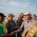 An outdoor gathering at what appears to be an airfield or aviation event, where a civilian elderly man in a white cap and gray polo shirt is shaking hands with a person wearing a high-visibility safety vest. Several military personnel in camouflage uniforms are present in the background. The setting suggests an official meet-and-greet or reception event at an airshow or military aviation function.
