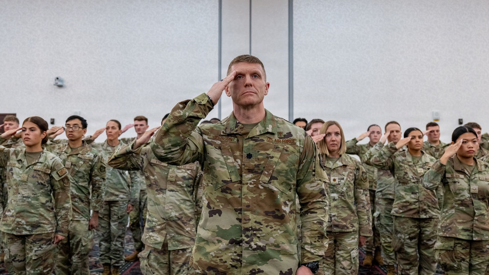 A large group of U.S. Air Force personnel in OCP (Operational Camouflage Pattern) uniforms are rendering a salute during what appears to be a formal military ceremony or formation. A central figure, prominently positioned in the foreground, displays a name tape reading 'JOHNSON' and a rank insignia consistent with a Lieutenant Colonel (oak leaf cluster), with 'U.S. AIR FORCE' tape visible on the chest. The setting appears to be an indoor venue such as a conference hall or event center.