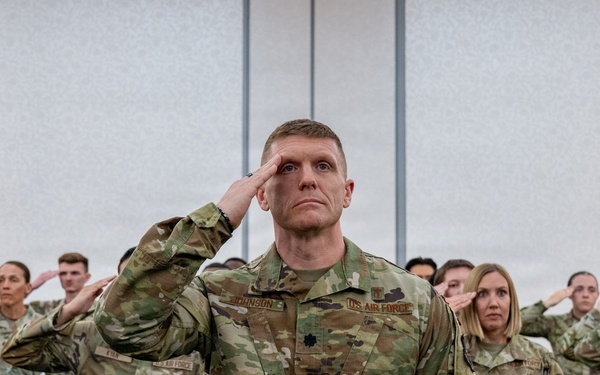 A large group of U.S. Air Force personnel in OCP (Operational Camouflage Pattern) uniforms are rendering a salute during what appears to be a formal military ceremony or formation. A central figure, prominently positioned in the foreground, displays a name tape reading 'JOHNSON' and a rank insignia consistent with a Lieutenant Colonel (oak leaf cluster), with 'U.S. AIR FORCE' tape visible on the chest. The setting appears to be an indoor venue such as a conference hall or event center.