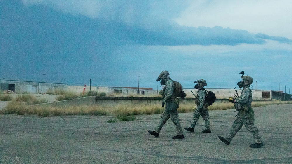 Three military personnel in full combat gear, including gas masks and helmets with mounted devices, are walking across a cracked tarmac or pavement surface. The setting appears to be an abandoned or derelict military installation with low concrete buildings visible in the background. The scene is captured under overcast, stormy-looking skies suggesting either dusk or an approaching storm.