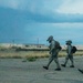 Three military personnel in full combat gear, including gas masks and helmets with mounted devices, are walking across a cracked tarmac or pavement surface. The setting appears to be an abandoned or derelict military installation with low concrete buildings visible in the background. The scene is captured under overcast, stormy-looking skies suggesting either dusk or an approaching storm.