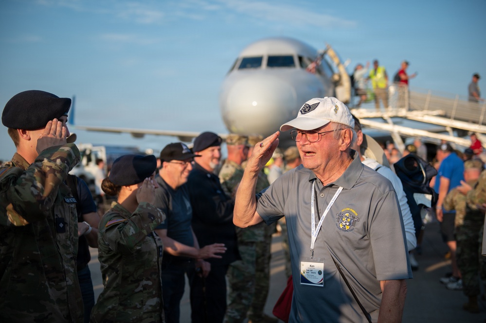 An elderly veteran named 'Ken' (visible on his name badge) is saluting on an airport tarmac, surrounded by active-duty military personnel in camouflage uniforms. The scene appears to be a honor flight or veteran recognition event, with a commercial aircraft visible in the background and people descending the boarding stairs. The emotional moment captures an exchange of salutes between the veteran and younger service members.