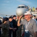 An elderly veteran named 'Ken' (visible on his name badge) is saluting on an airport tarmac, surrounded by active-duty military personnel in camouflage uniforms. The scene appears to be a honor flight or veteran recognition event, with a commercial aircraft visible in the background and people descending the boarding stairs. The emotional moment captures an exchange of salutes between the veteran and younger service members.