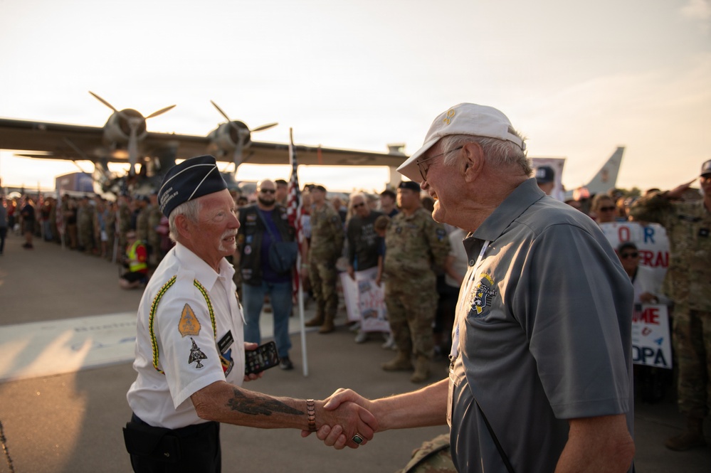 Two elderly men are shaking hands on what appears to be an airfield tarmac during a veteran or military honor event. One man wears a military-style white uniform shirt with patches and a garrison cap, while the other wears a gray polo shirt and white baseball cap. A large crowd of military personnel, civilians, and supporters lines the background, with a vintage multi-engine aircraft visible behind them.