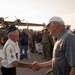 Two elderly men are shaking hands on what appears to be an airfield tarmac during a veteran or military honor event. One man wears a military-style white uniform shirt with patches and a garrison cap, while the other wears a gray polo shirt and white baseball cap. A large crowd of military personnel, civilians, and supporters lines the background, with a vintage multi-engine aircraft visible behind them.