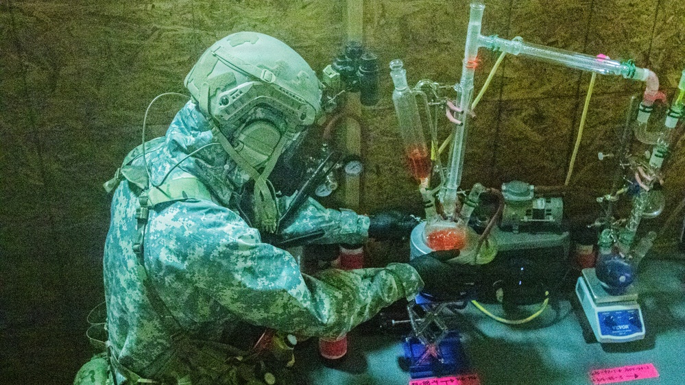 A person wearing full military-style CBRN (Chemical, Biological, Radiological, Nuclear) protective gear and a tactical helmet with respirator is examining chemical laboratory equipment. The setting appears to be a makeshift or field laboratory environment with plywood walls. This appears consistent with law enforcement or military documentation of a clandestine drug laboratory raid or training exercise.