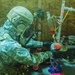 A person wearing full military-style CBRN (Chemical, Biological, Radiological, Nuclear) protective gear and a tactical helmet with respirator is examining chemical laboratory equipment. The setting appears to be a makeshift or field laboratory environment with plywood walls. This appears consistent with law enforcement or military documentation of a clandestine drug laboratory raid or training exercise.
