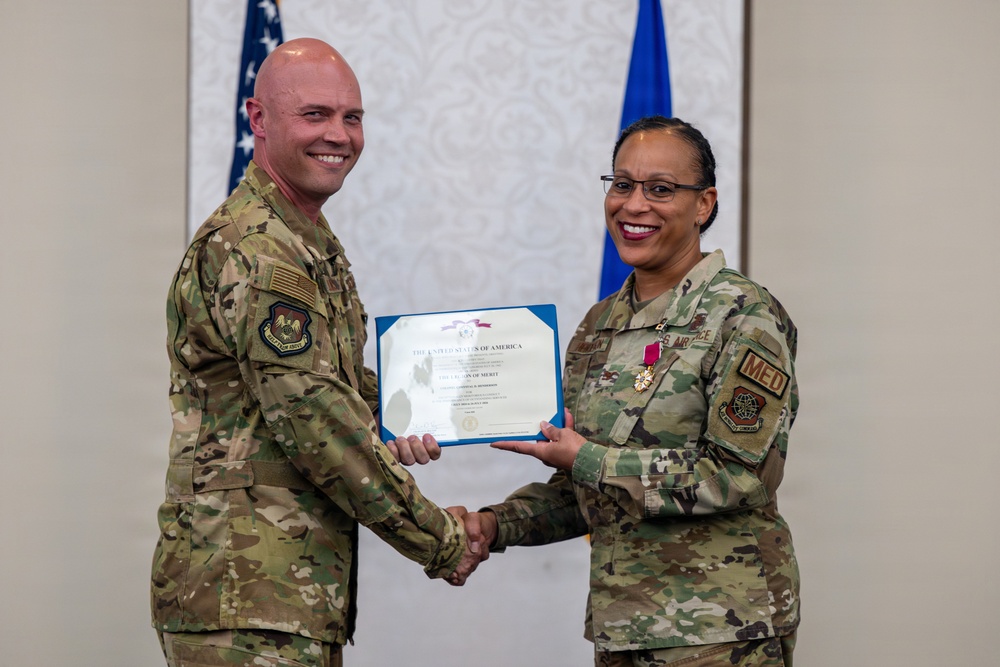 Two U.S. Air Force personnel in OCP (Operational Camouflage Pattern) uniforms are posing together while jointly holding a Legion of Merit certificate. The female service member on the right is wearing the Legion of Merit medal on her uniform, indicating she is the award recipient. The ceremony appears to be taking place in a formal indoor setting with U.S. and Air Force flags in the background.