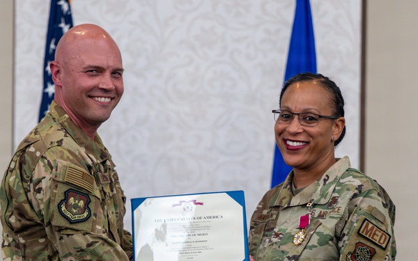 Two U.S. Air Force personnel in OCP (Operational Camouflage Pattern) uniforms are posing together while jointly holding a Legion of Merit certificate. The female service member on the right is wearing the Legion of Merit medal on her uniform, indicating she is the award recipient. The ceremony appears to be taking place in a formal indoor setting with U.S. and Air Force flags in the background.