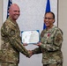 Two U.S. Air Force personnel in OCP (Operational Camouflage Pattern) uniforms are posing together while jointly holding a Legion of Merit certificate. The female service member on the right is wearing the Legion of Merit medal on her uniform, indicating she is the award recipient. The ceremony appears to be taking place in a formal indoor setting with U.S. and Air Force flags in the background.