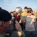 An elderly Vietnam War Air Force veteran is saluting during what appears to be an honor flight event at an airport tarmac. He is wearing a black cap reading 'Vietnam Veteran Air Force' and an Under Armour 'Freedom' shirt, being greeted by military personnel and civilians. A commercial aircraft is visible in the background along with a crowd of attendees.