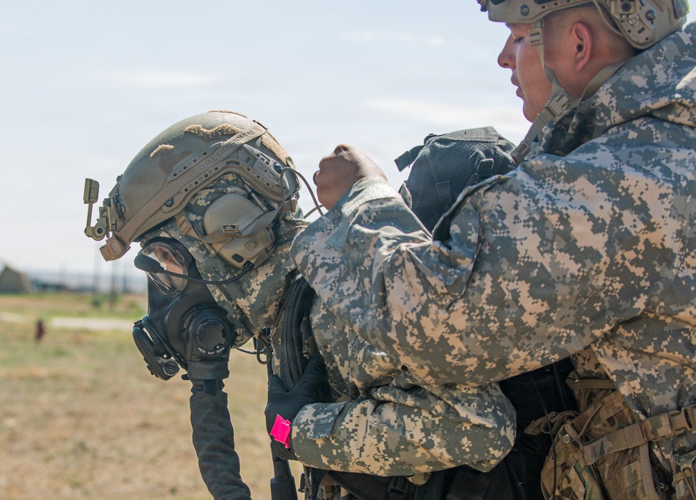Two military personnel in digital camouflage uniforms are shown in an outdoor field environment. One soldier wearing a tactical helmet with ear protection and a gas mask has their equipment being adjusted or checked by a second soldier. The setting appears to be a training exercise or pre-mission preparation.