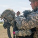 Two military personnel in digital camouflage uniforms are shown in an outdoor field environment. One soldier wearing a tactical helmet with ear protection and a gas mask has their equipment being adjusted or checked by a second soldier. The setting appears to be a training exercise or pre-mission preparation.